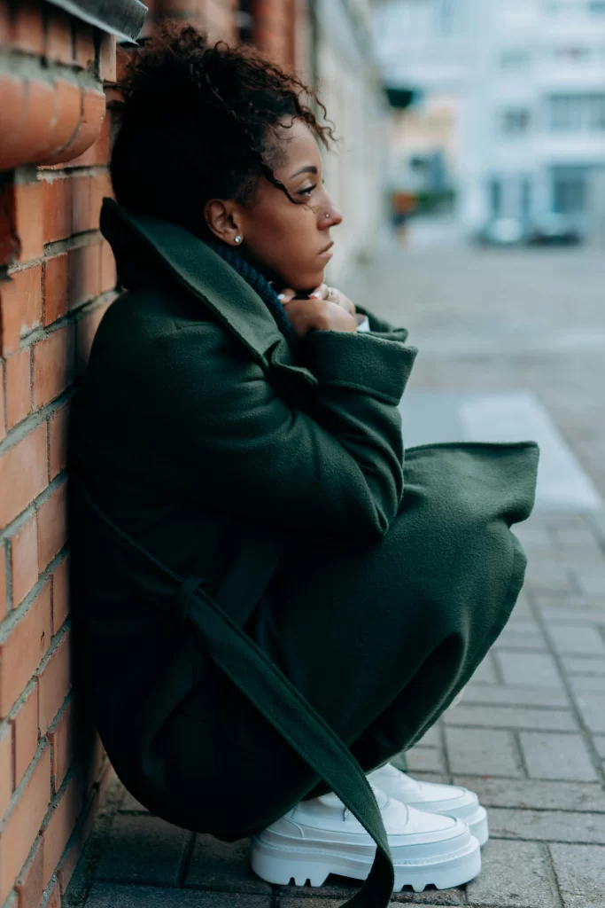 A woman in a trench coat leans against a brick wall, looking contemplative on a city sidewalk.