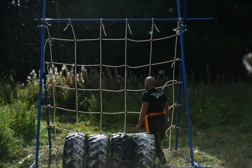 Man training on an outdoor obstacle course, featuring a climbing net and tires.