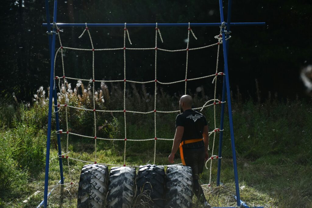 Man training on an outdoor obstacle course, featuring a climbing net and tires.
